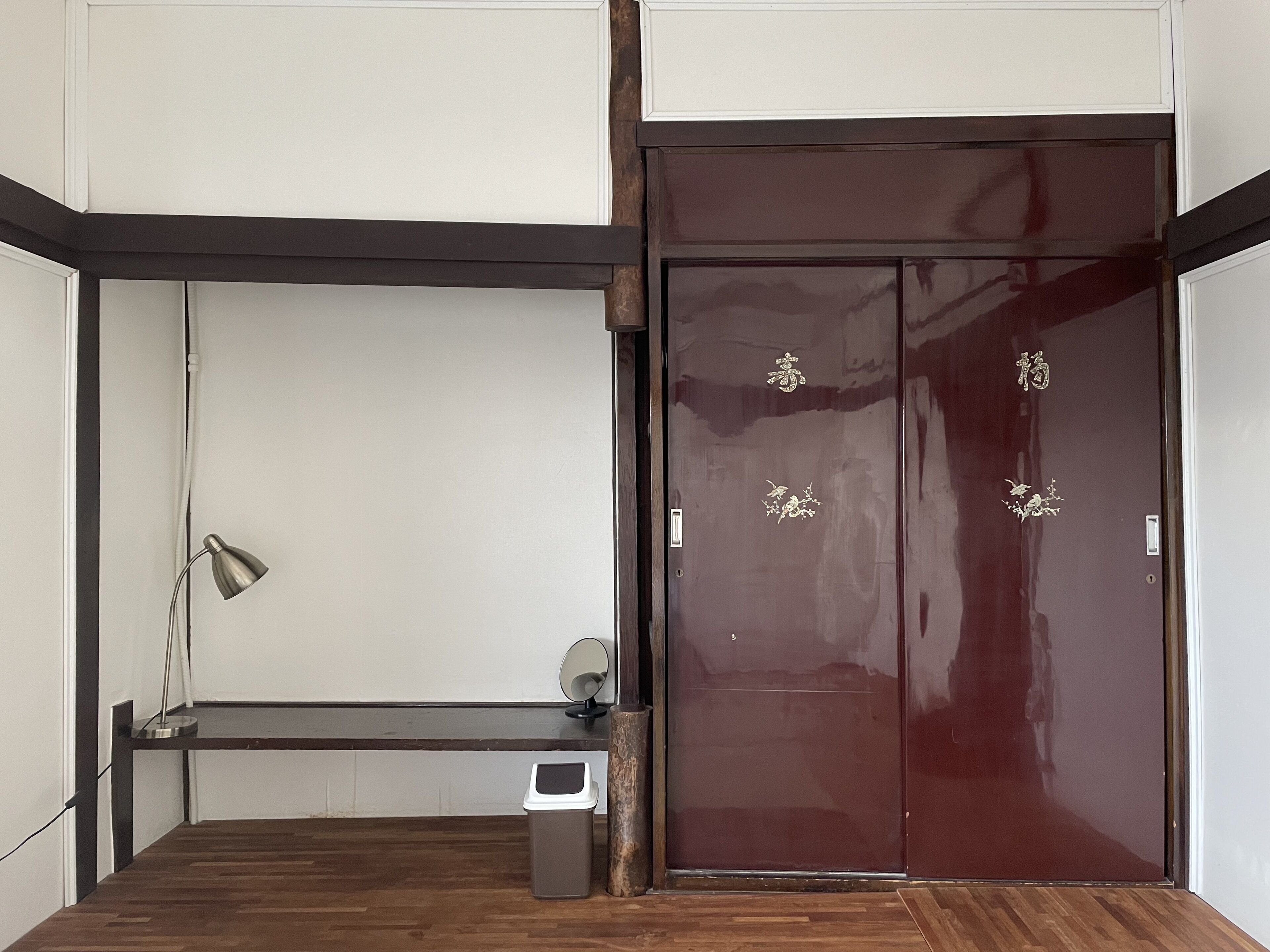 A room with a wooden floor, a desk with a lamp, a trash bin, and a large dark wooden door with decorative gold accents.