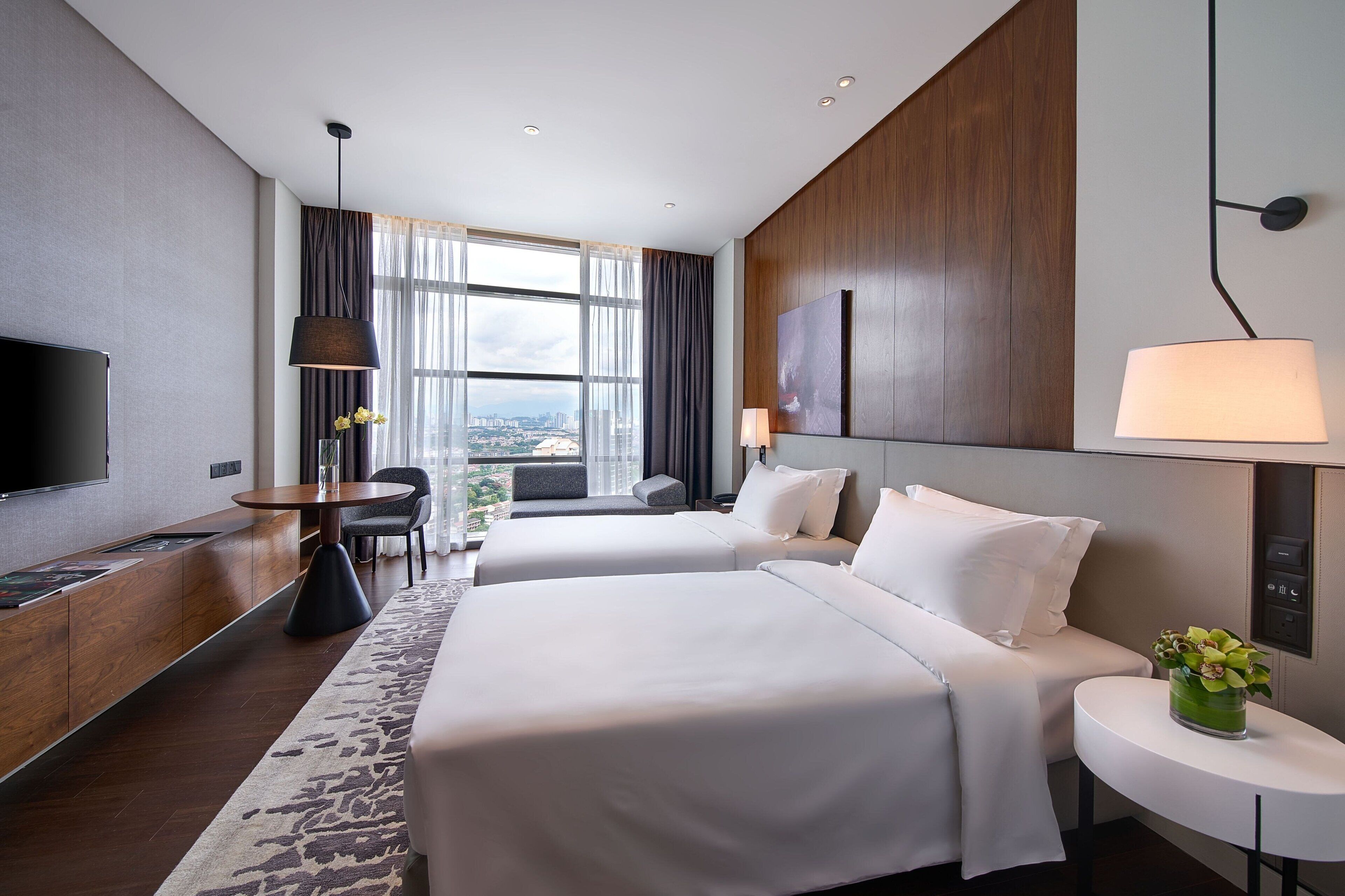 A modern hotel room with two beds, a flat-screen TV, a dining area, and a large window offering a city view.