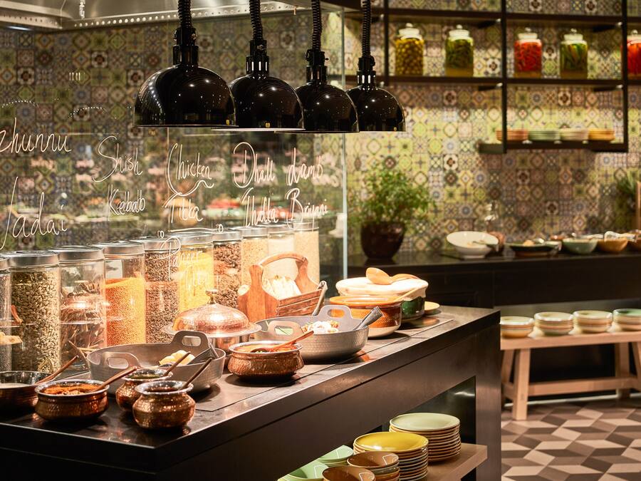 Un área de bufé con varios recipientes y platos de comida, una barra con platos apilados y una pared de azulejos con diseño al fondo.