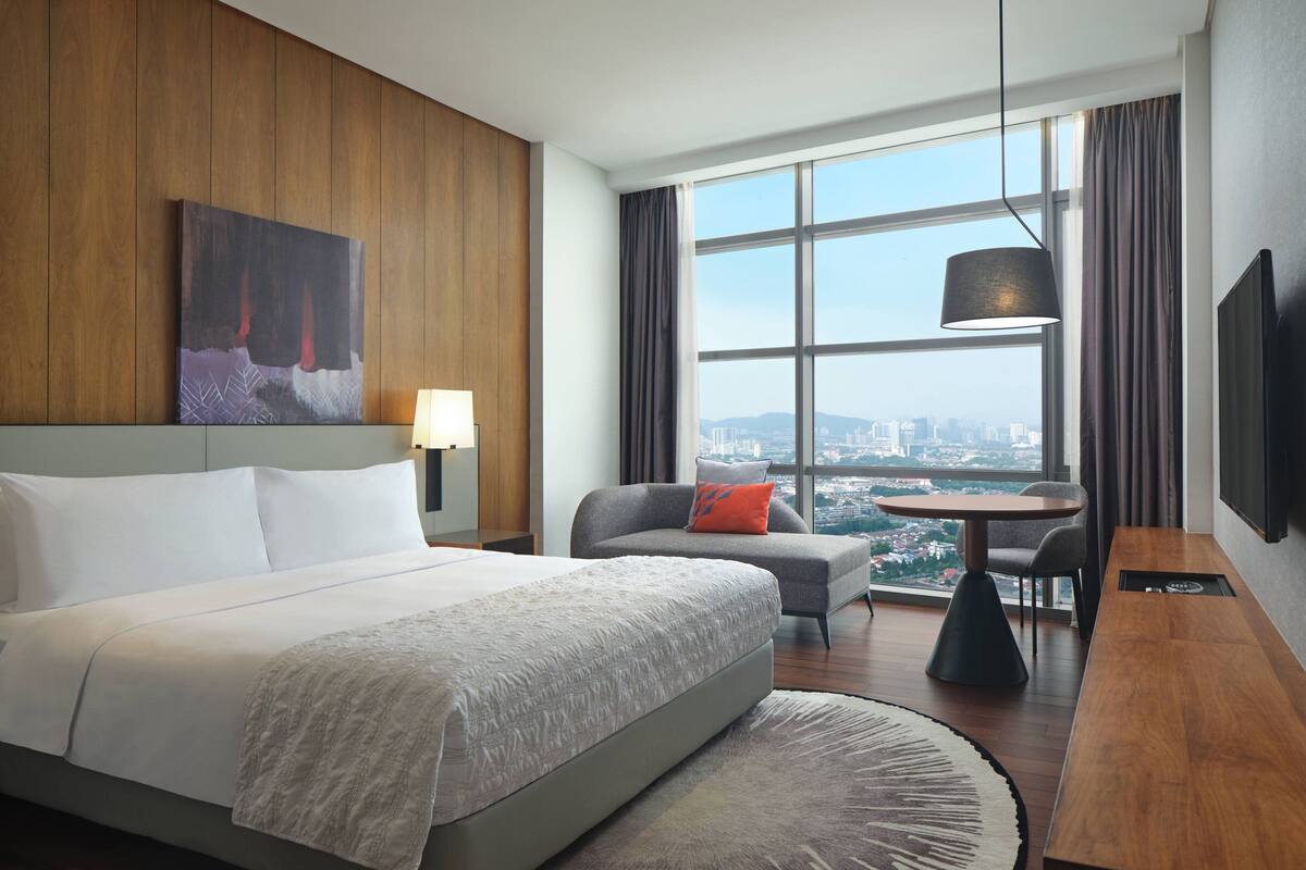 Ein modernes Hotelzimmer mit einem großen Bett, einer Sitzecke, einem Esstisch und Blick auf die Stadt durch große Fenster.