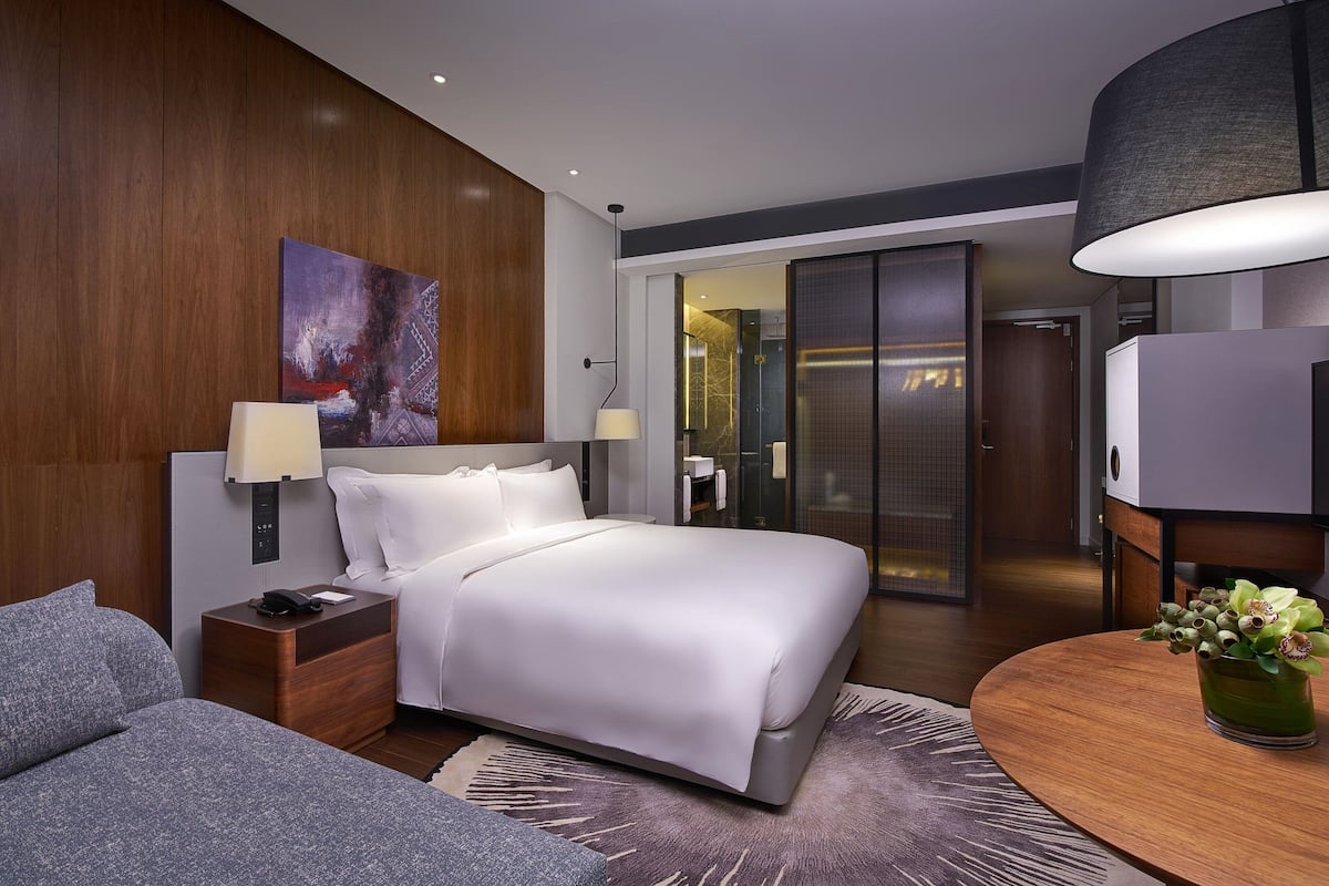 Ein modernes Hotelzimmer mit einem großen Bett, Nachttischlampen, einem kleinen Tisch mit Blumenvase und einem Gemälde an der Wand.