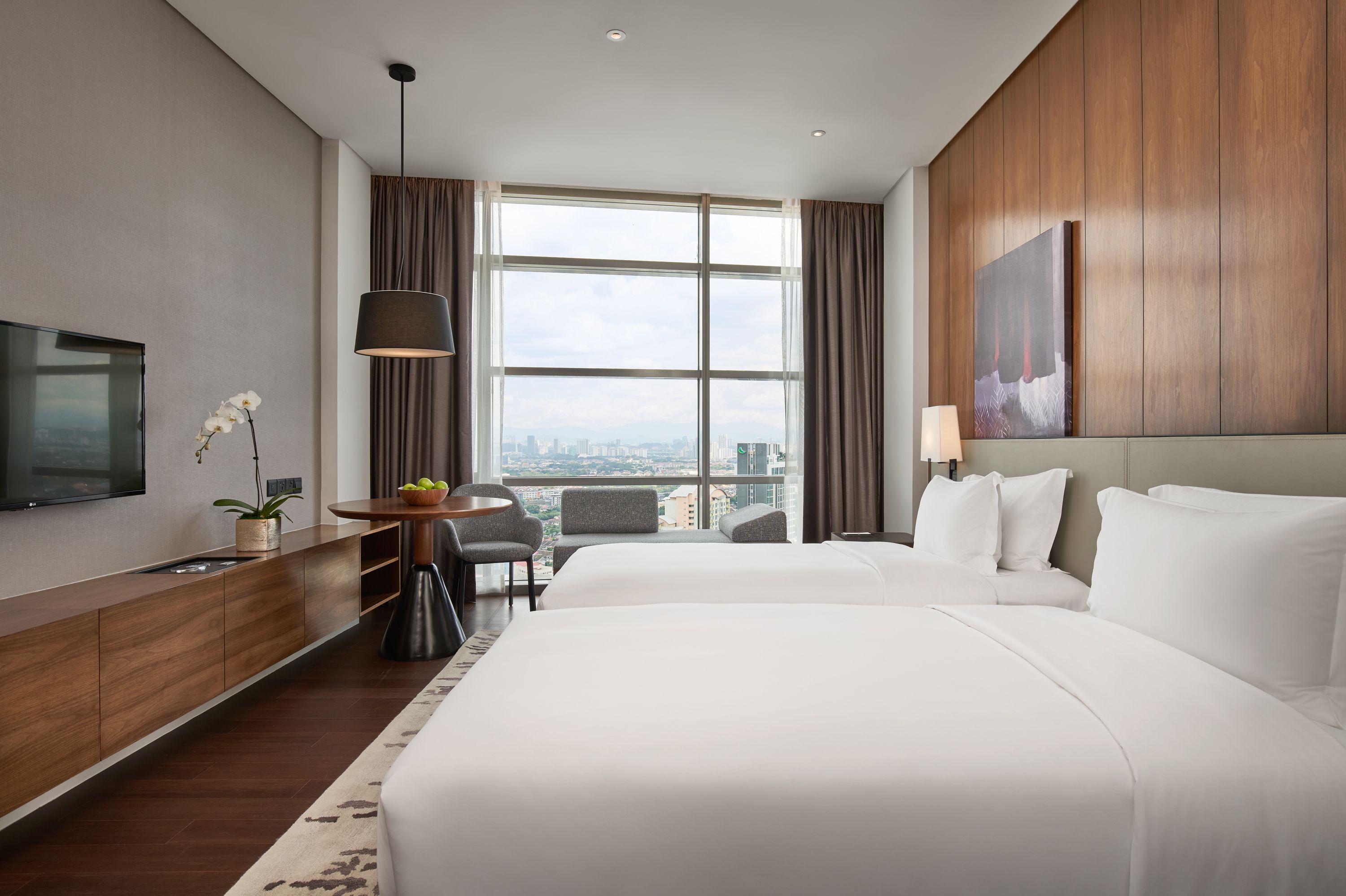 A hotel room with a large bed, a TV, a desk with a lamp, a chair, and a view of the city through large windows.