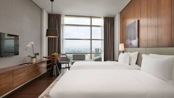 Quarto de hotel com uma cama grande, televisão, escrivaninha com luminária, cadeira e vista da cidade através de amplas janelas.
