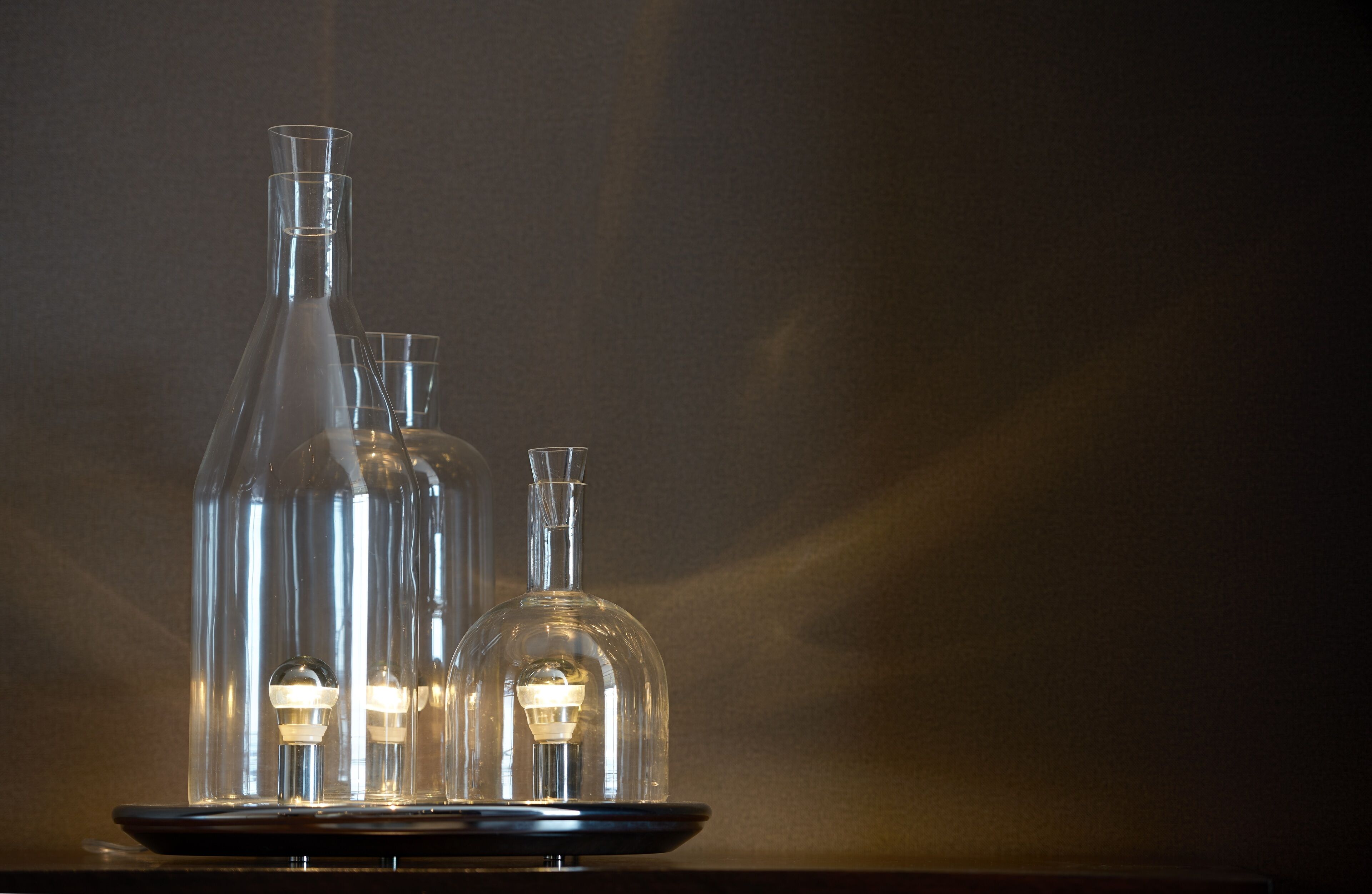Tres botellas de vidrio con bombillas en su interior, sobre una bandeja negra y un fondo oscuro.