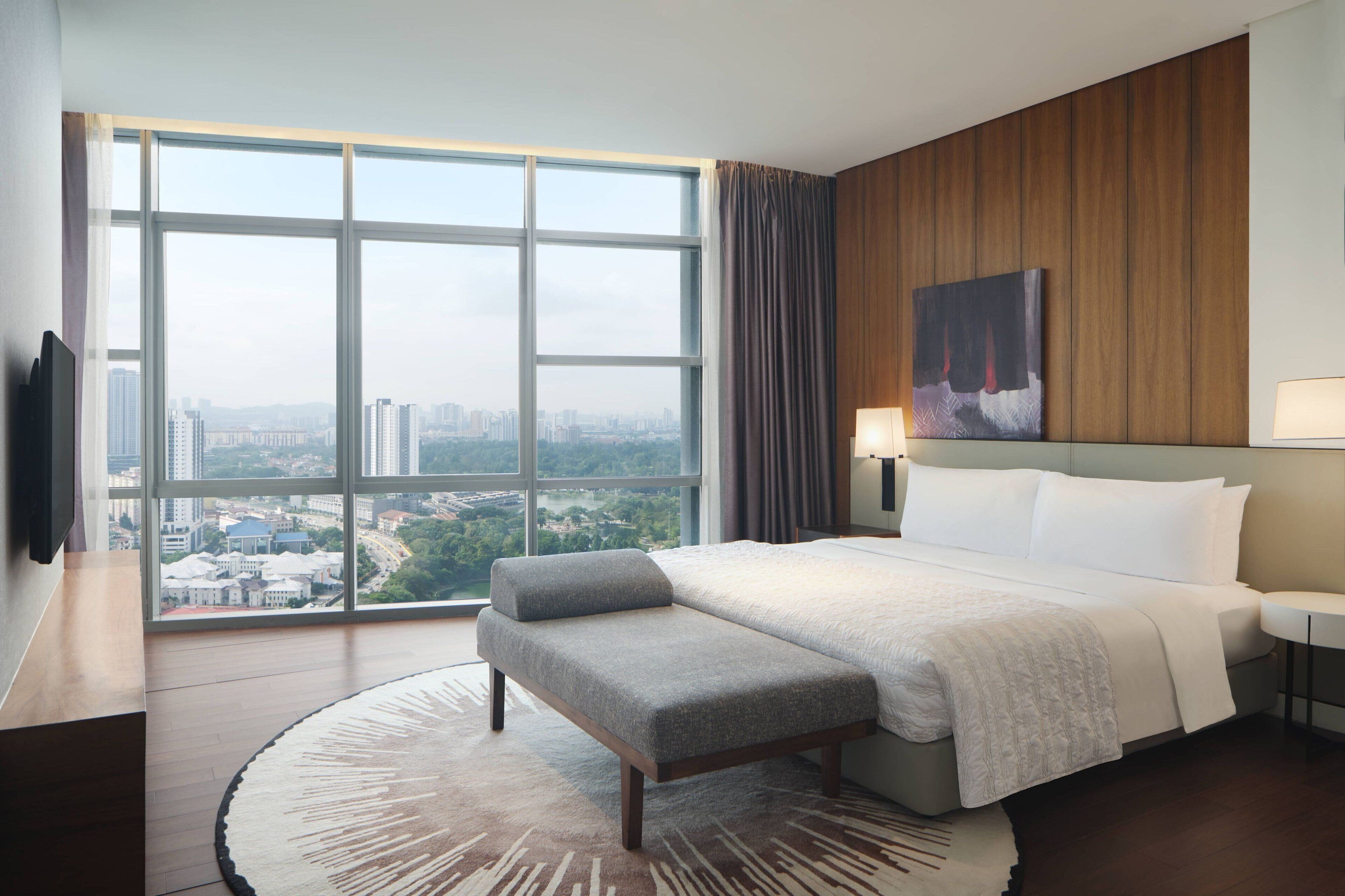 A hotel room with a large window, a bed with a grey bench, a wooden panel wall, and a painting.