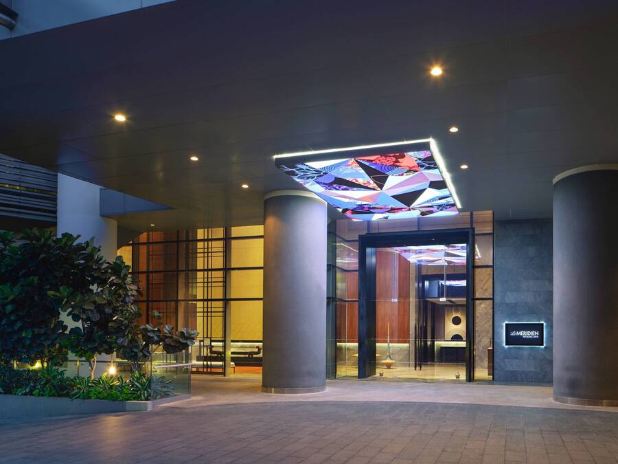 A modern hotel entrance with a geometric light installation above the doors, large columns, and a well-lit reception area.