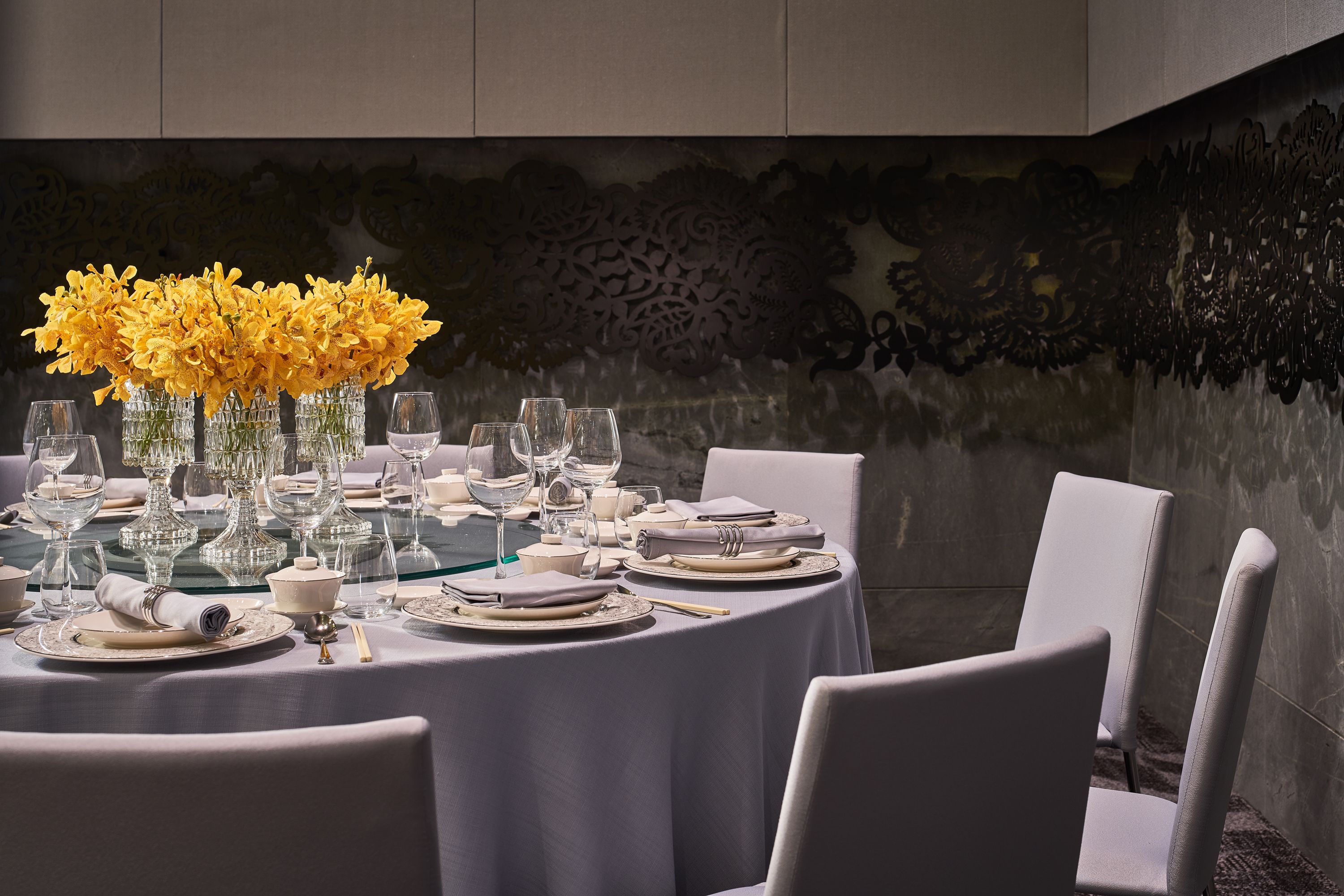 Une table de salle à manger ronde, recouverte d’une nappe blanche, agrémentée de verres et de fleurs jaunes disposées dans des vases.