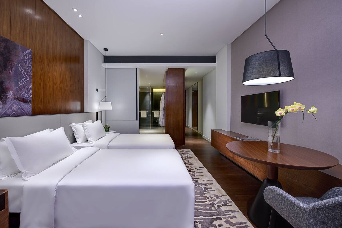 Ein modernes Hotelzimmer mit zwei Betten, einem Schreibtisch, einem Sessel, einem großen Gemälde, einem Fernseher und einer Vase mit Blumen.