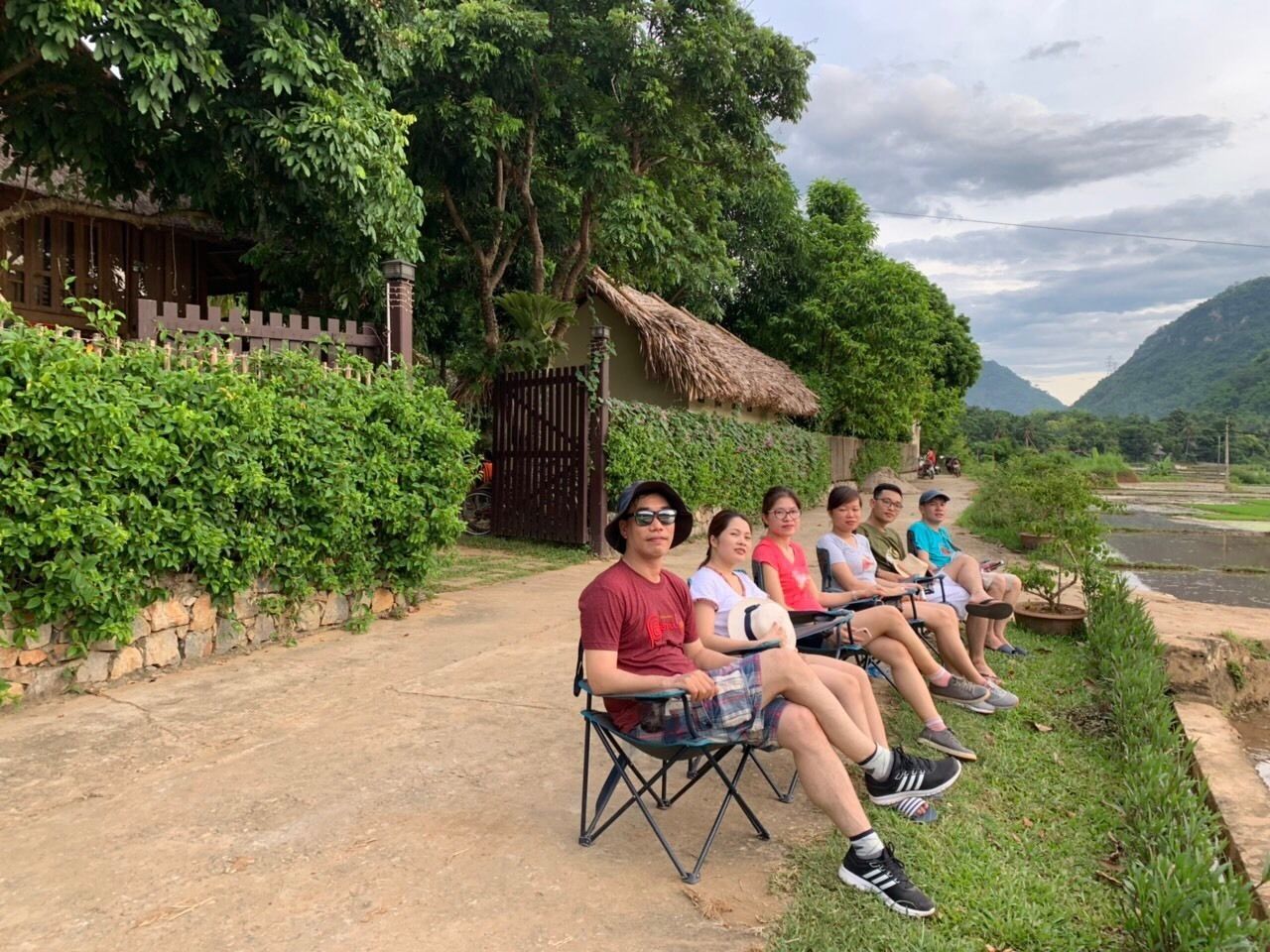 A group of people seated on folding chairs along a dirt path beside a thatched-roof structure.