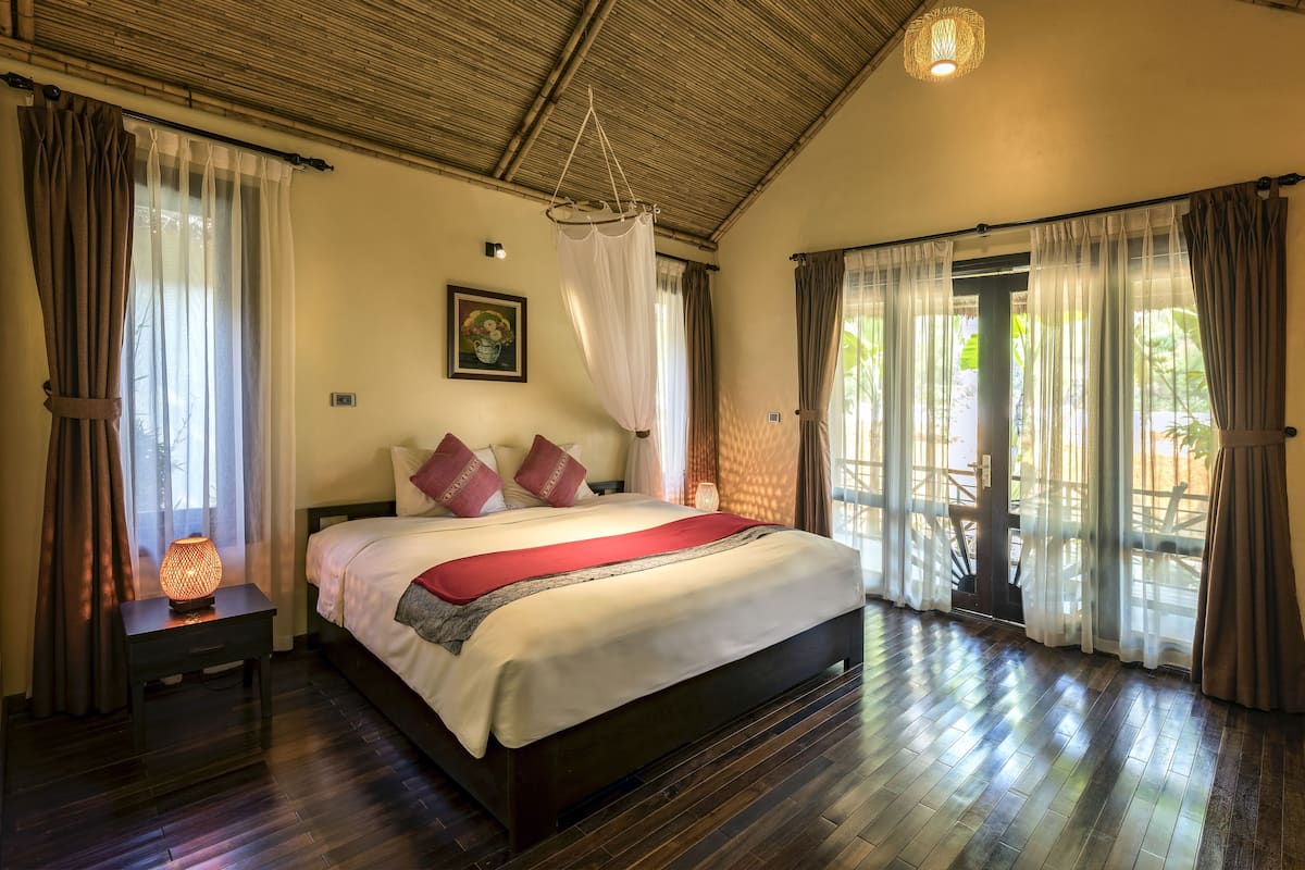 Een slaapkamer met een groot bed, houten vloeren, een hanglamp en uitzicht door een glazen deur.