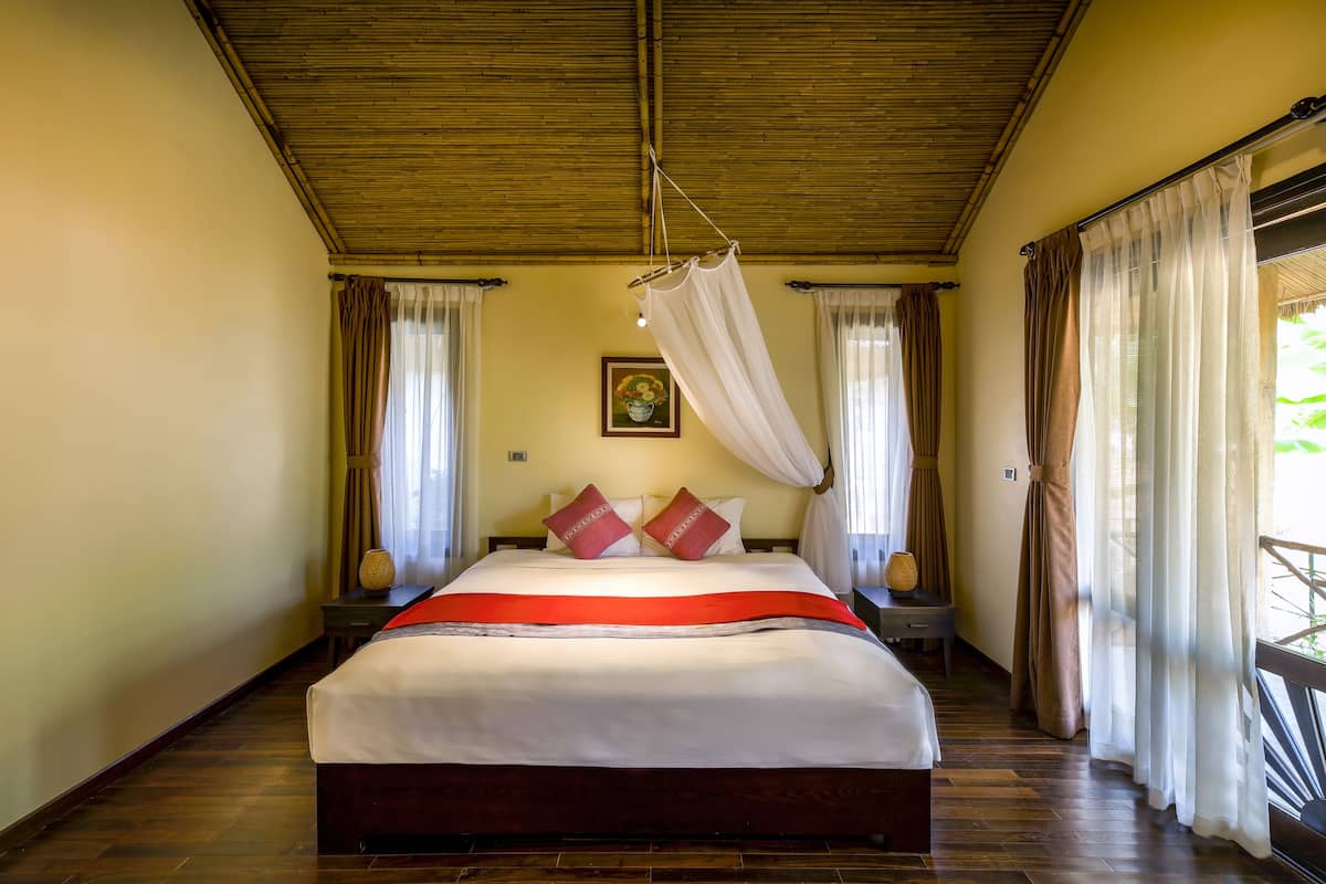 Een slaapkamer met een bed, houten vloer, gordijnen en een hangend muggengaas.