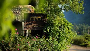 Une pancarte rustique sur laquelle on peut lire « Village de Mau Cha », entourée d’une végétation luxuriante et de fleurs roses.