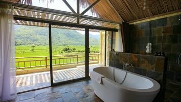 A bathroom with a large bathtub, a stone wall, and a view of a green landscape.