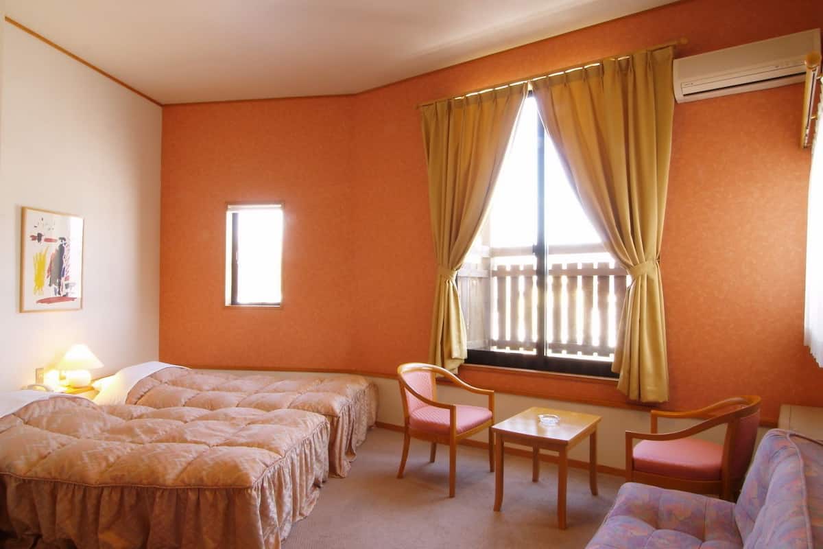 A hotel room with a bed, a small table, two chairs, a sofa, a window with curtains, a wall-mounted air conditioner, and a framed picture on the wall.