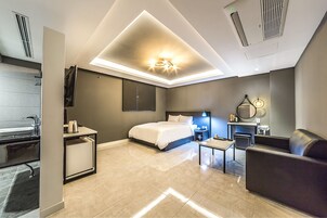 A modern hotel room with a bed, a desk, a chair, a sofa, a mirror, and a bathroom with a sink and toilet.