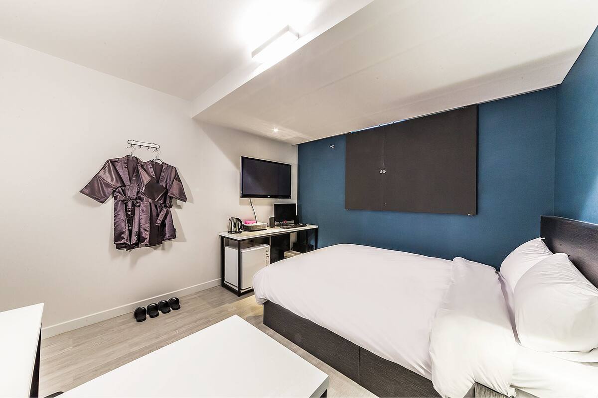 A modern hotel room with a large bed, a desk with a flat-screen TV, a small cabinet, and a wall-mounted TV.