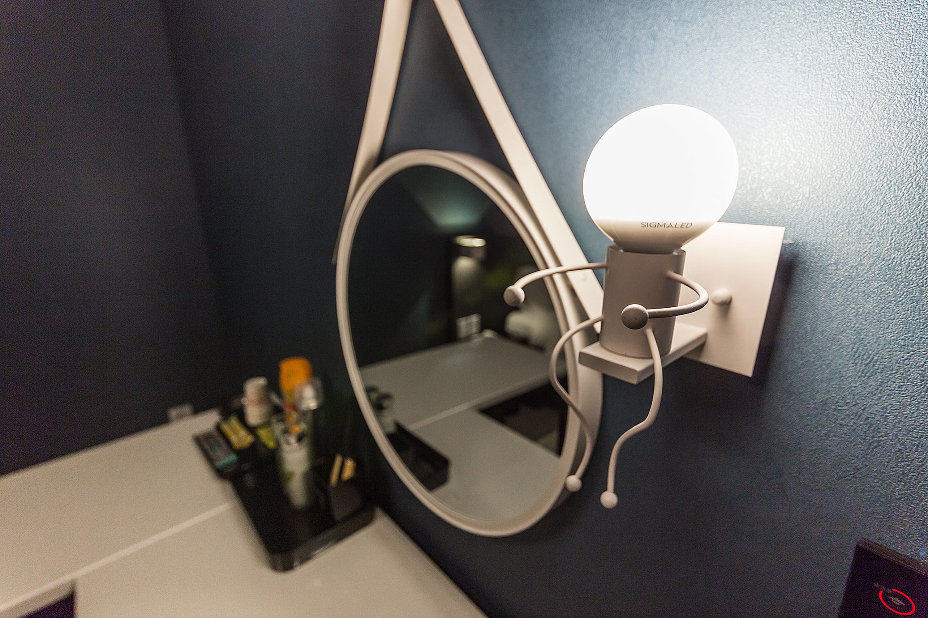 Un baño con espejo instalado en la pared, iluminación y una encimera blanca con artículos de tocador.