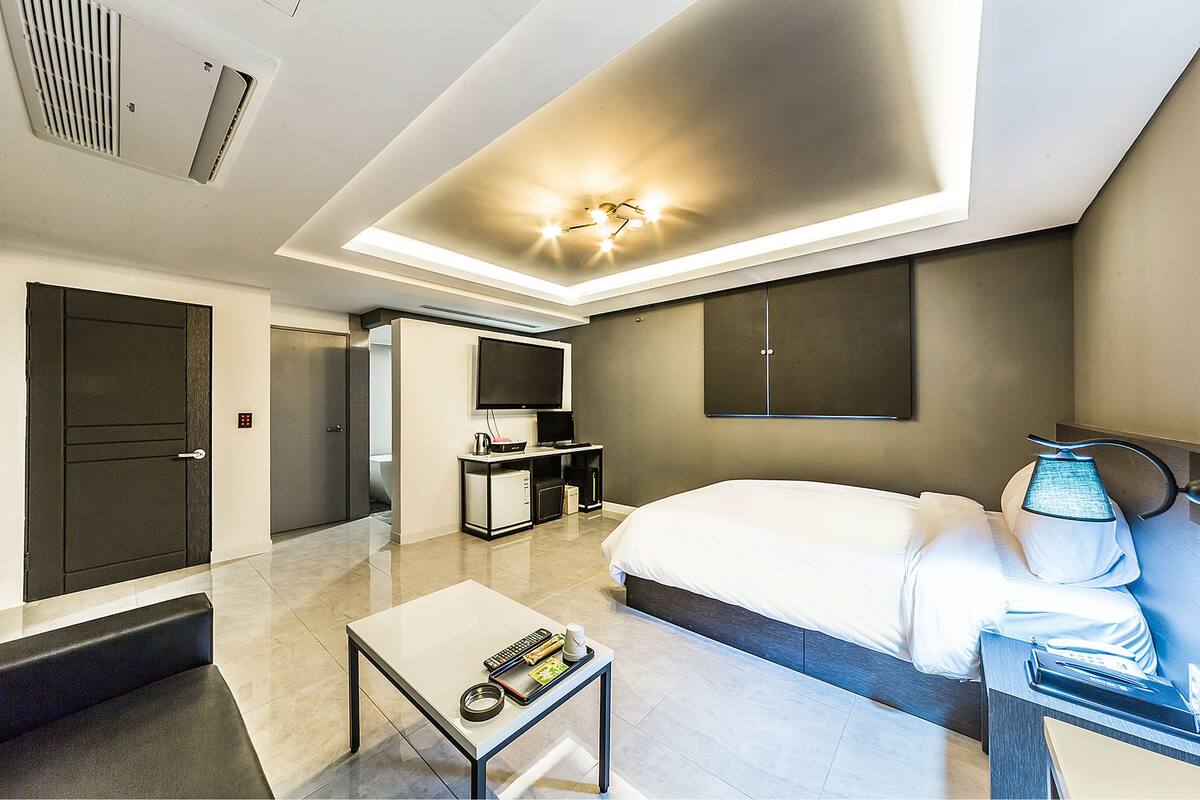 A modern hotel room with a bed, a desk, a coffee table, a TV, and a minibar.