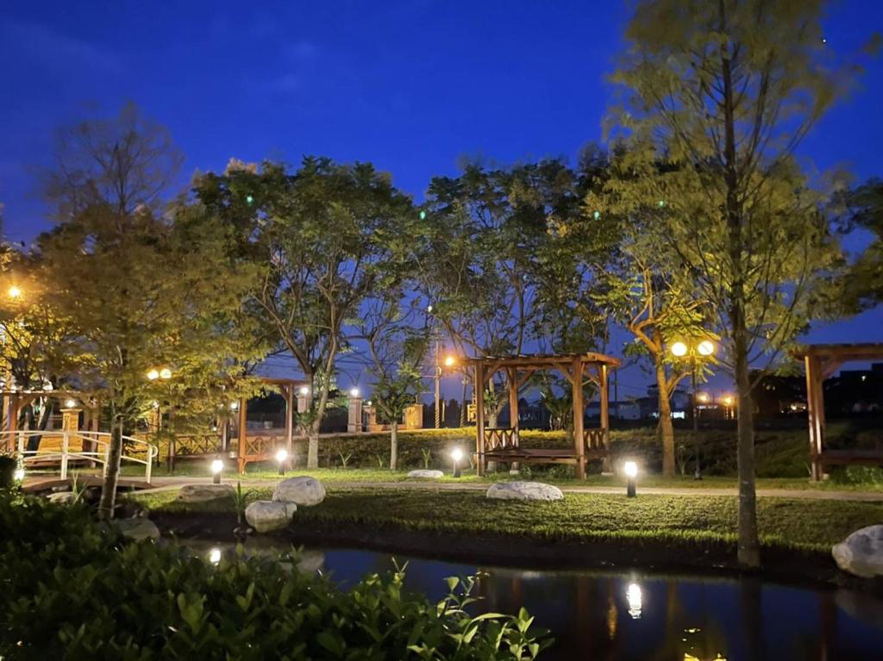 A well-lit park area with a pond, benches, and a gazebo.