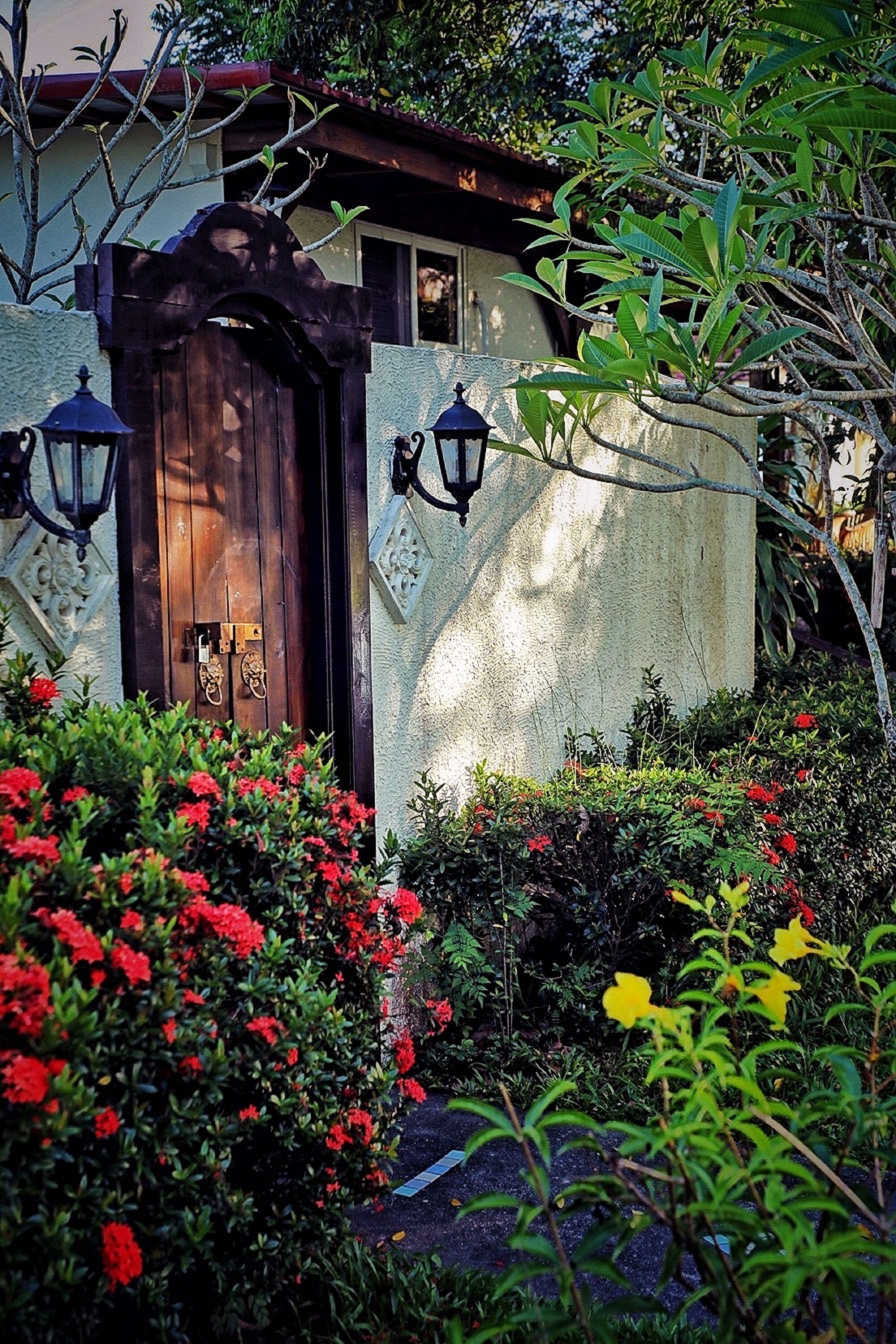 Uma casa com porta de madeira, duas lanternas fixadas na parede e um jardim com flores vermelhas e amarelas.