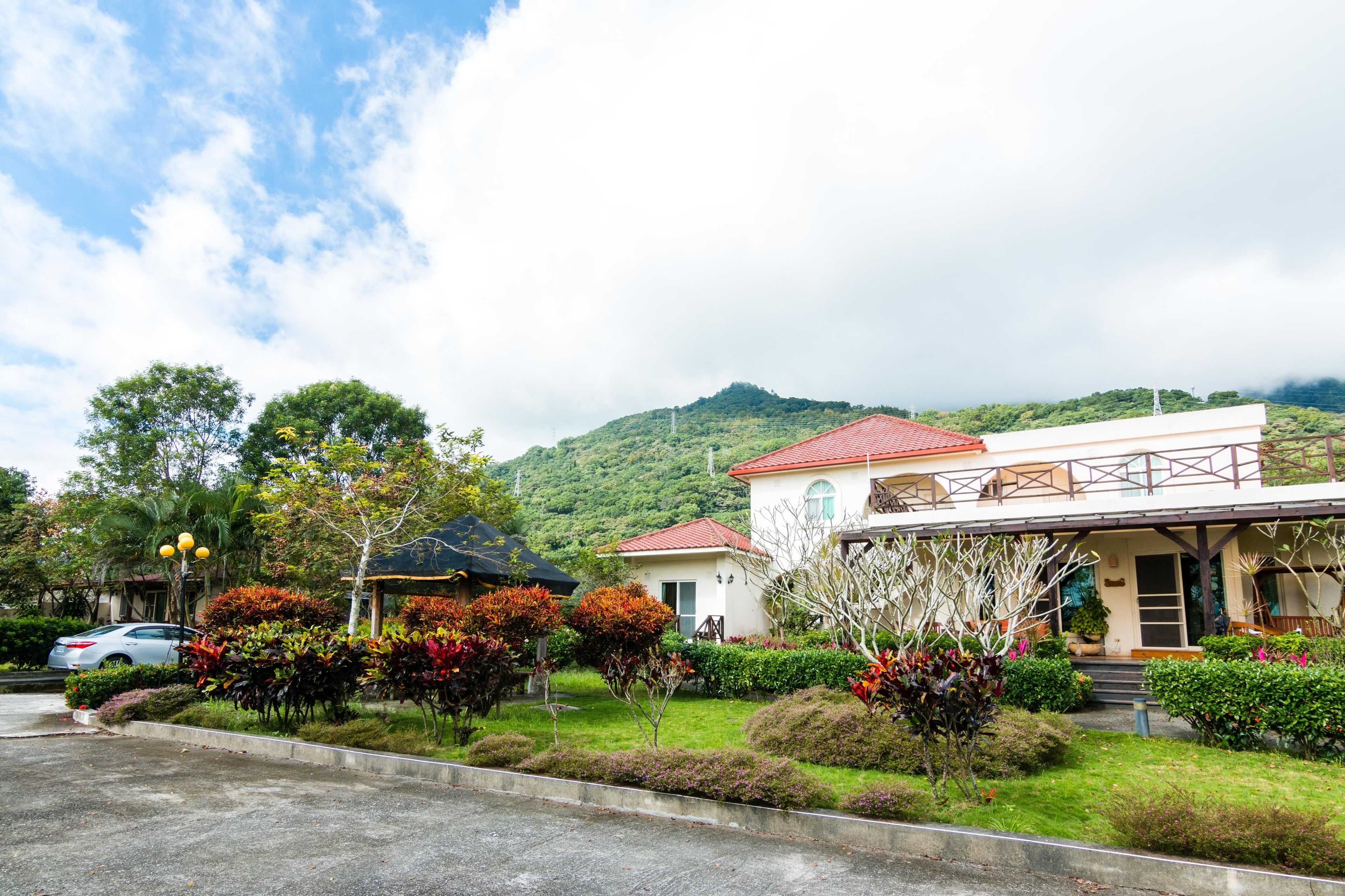 A two-story building with a red roof, surrounded by lush greenery and a mountainous backdrop.