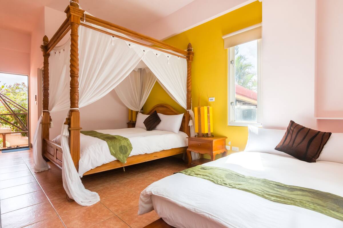 A four-poster bed with white bedding and a green throw, a bedside table with candles, and a window with a view of palm trees.