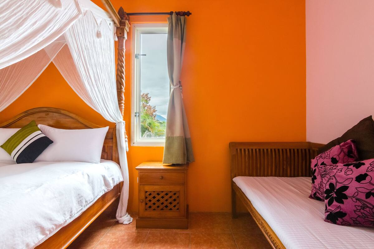 A bedroom with a wooden bed, a nightstand, a window with curtains, and a view of greenery outside.