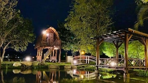 Un belvédère en bois avec un pont enjambant un étang, entouré d’arbres et illuminé par des lumières.