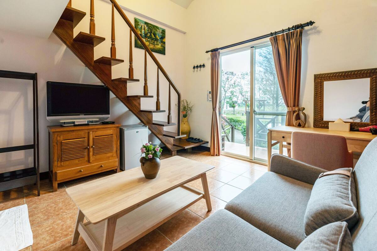 A living room with a wooden staircase, a flat-screen TV, a wooden coffee table with a vase, a grey sofa, and a sliding glass door leading to a balcony.