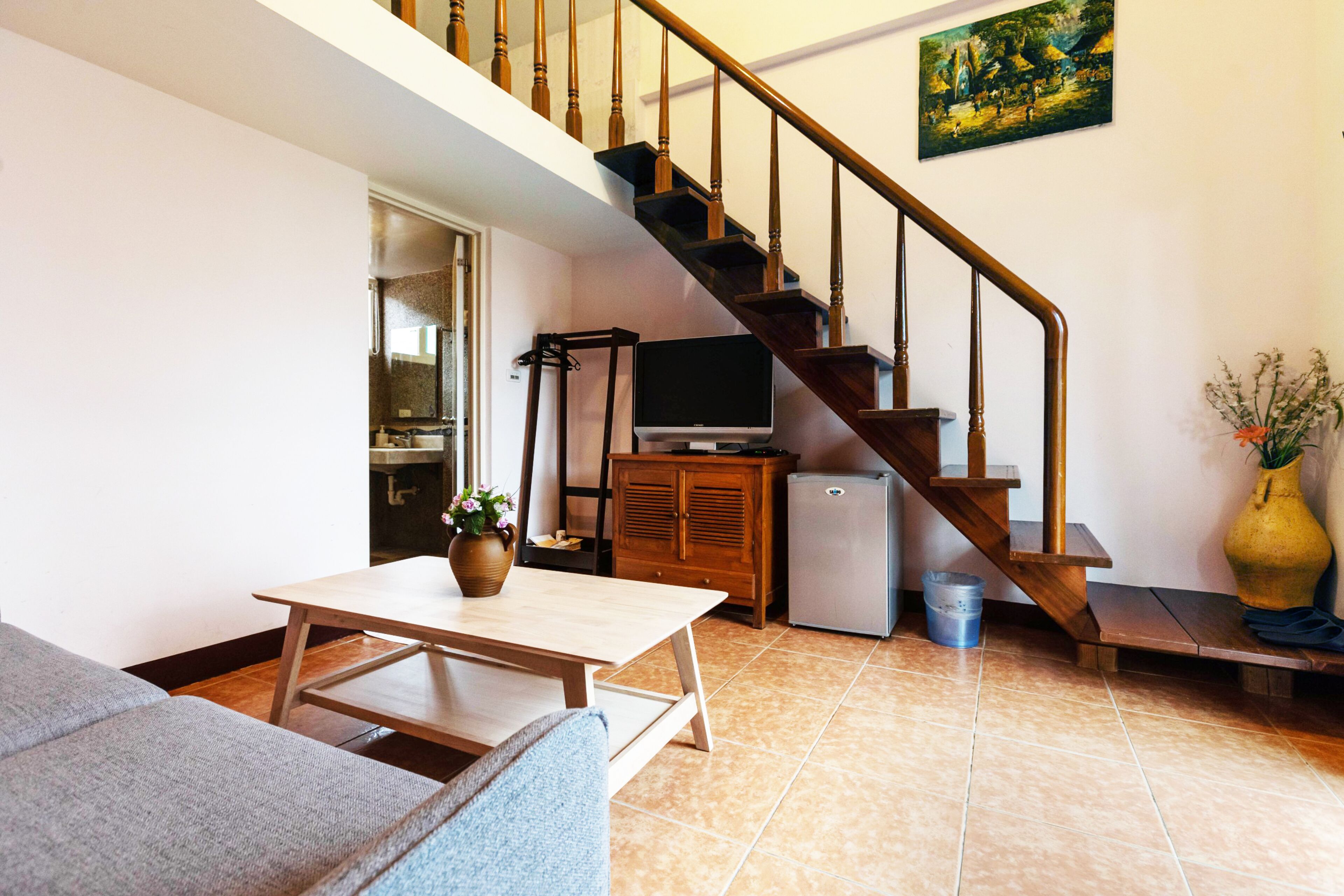 A living room with a staircase, a television, a small refrigerator, a sofa, and a table with a vase.