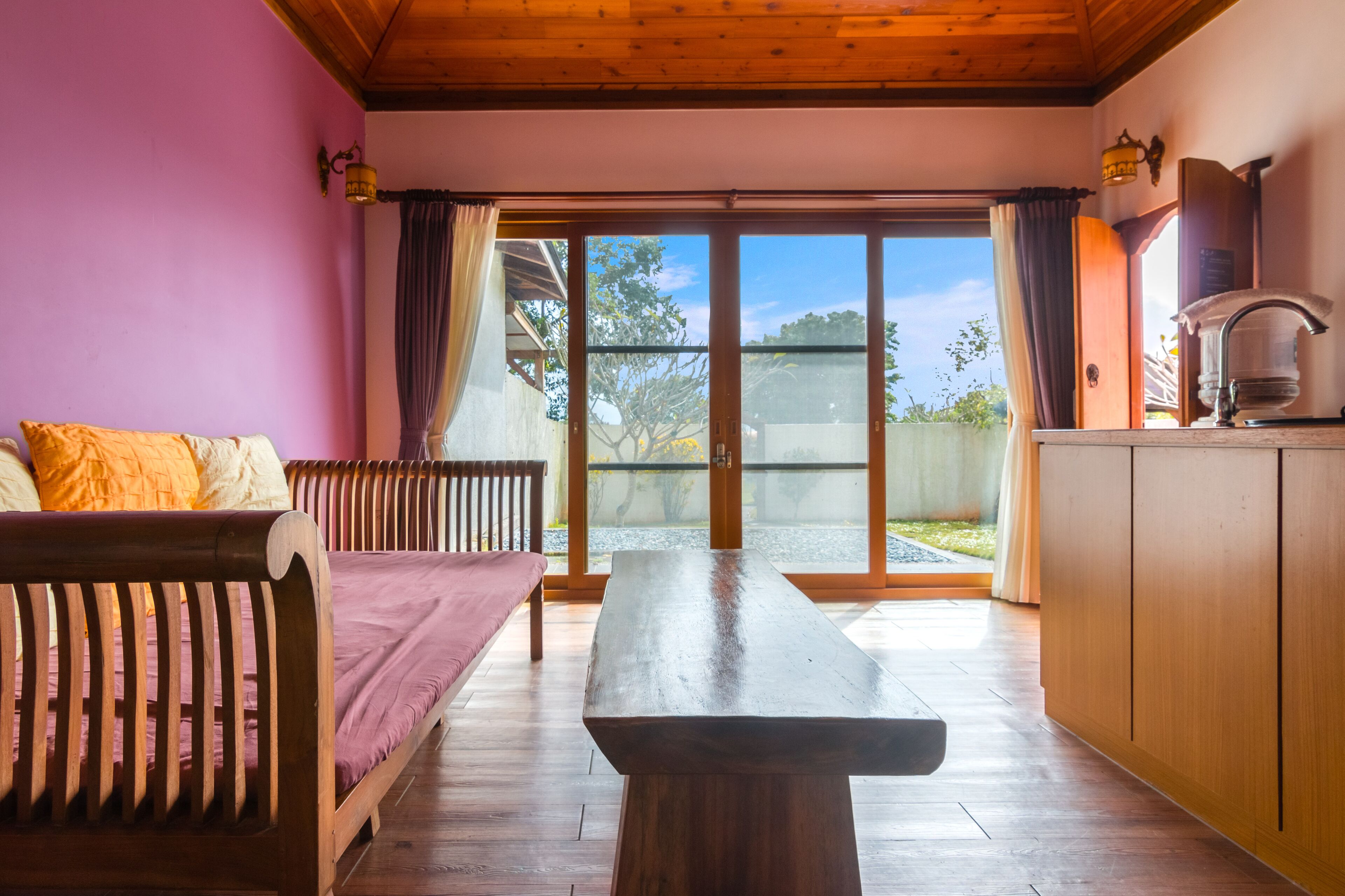Una habitación con un banco de madera, una mesa y un sofá con cojines. Hay un ventanal corredero que da a un espacio exterior.