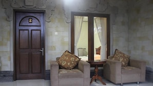 A room with a wooden door marked with the number 2, two armchairs with floral cushions, a small round table, and a mirror reflecting a bed with a nightstand. - Puri Anom Guest House (Denpasar)