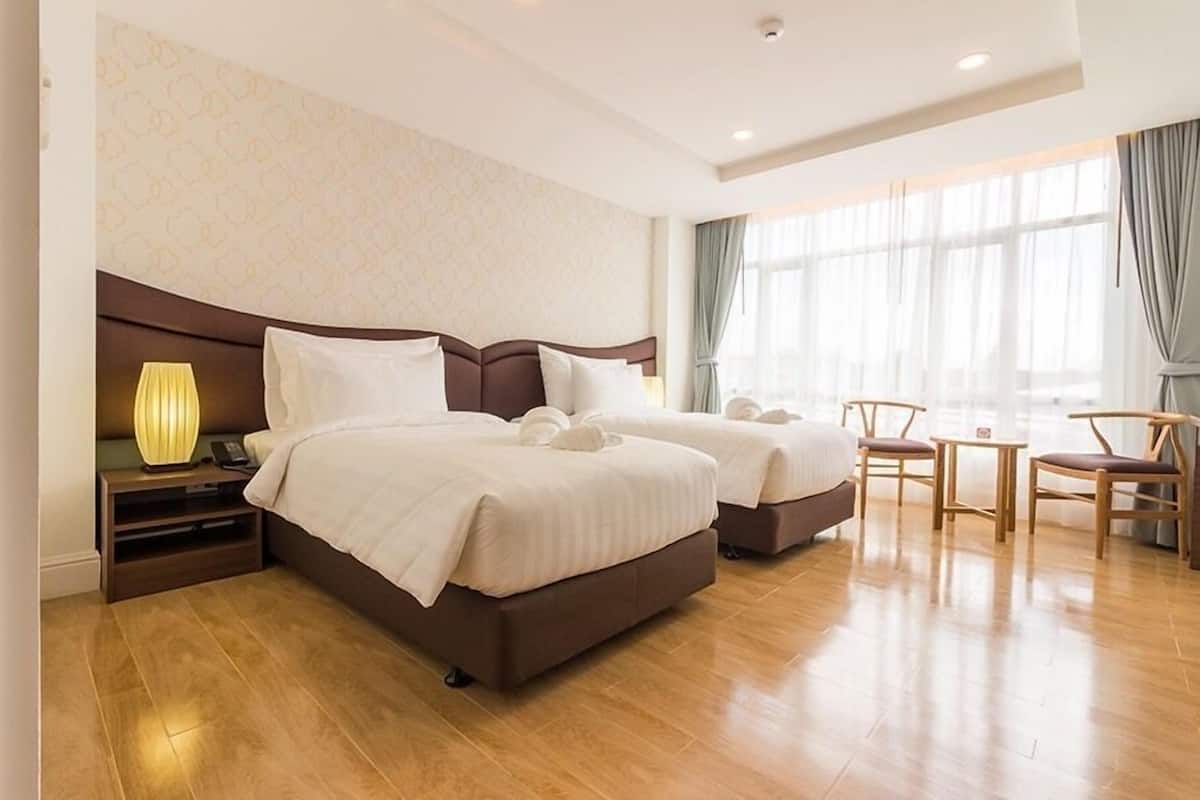 Habitación de hotel con dos camas, una mesita de noche con lámpara, una pequeña mesa con sillas y un ventanal con cortinas.