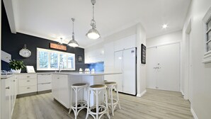 A modern kitchen with white cabinetry, a central island with stools, pendant lighting, and a clock on the wall. - Crusoe's Beach House Ocean Views (Cannonvale)