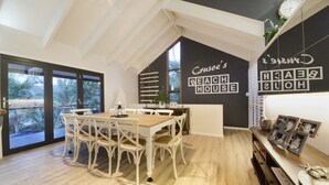 A dining area with a wooden table and white chairs, a black and white wall with 'Cruso's Beach House' text, and a view through a glass door. - Crusoe's Beach House Ocean Views (Cannonvale)