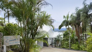 A sign welcoming to Canso's Beach House, surrounded by tropical vegetation and a view of the ocean. - Crusoe's Beach House Ocean Views (Cannonvale)