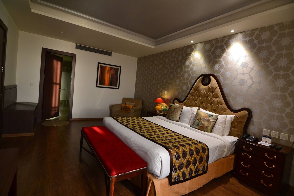 A hotel room with a large bed, a bench, a nightstand, and a door leading to another room.