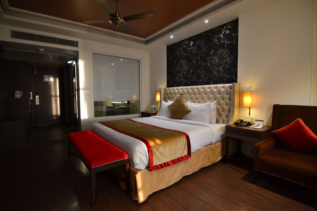 A hotel room with a large bed, a red bench, a wooden nightstand, and a red chair.