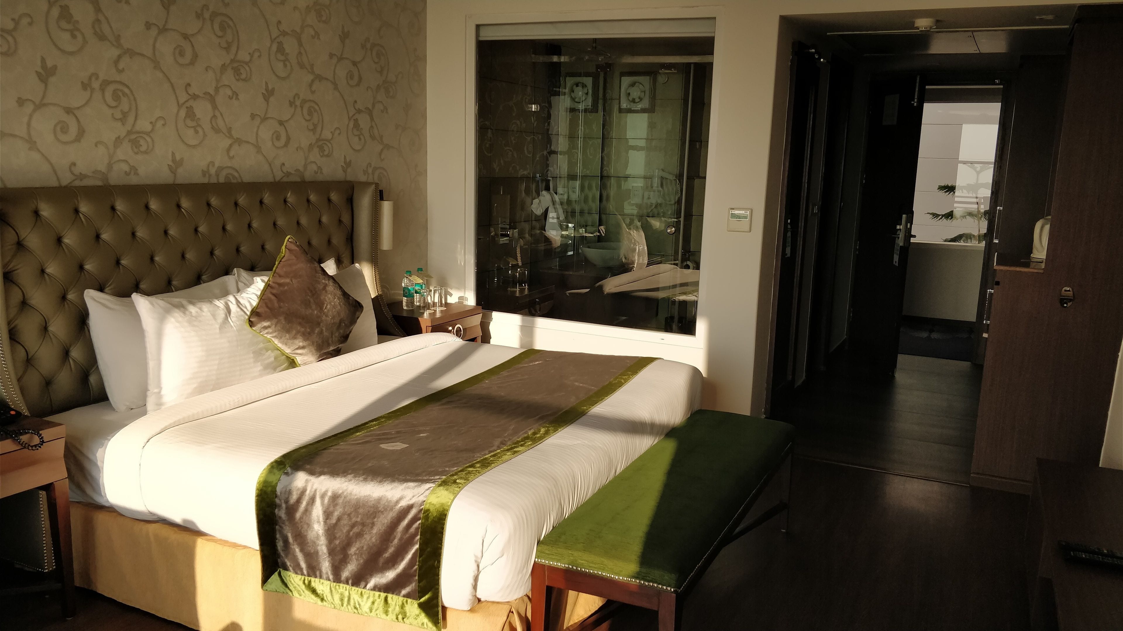 A well-appointed hotel room with a large bed, a bedside table with a lamp, and a bathroom visible through a glass door.