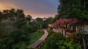 Un sendero conduce a una hilera de edificios de estilo tradicional con tejados rojos, rodeado de abundante vegetación y un telón de fondo montañoso.