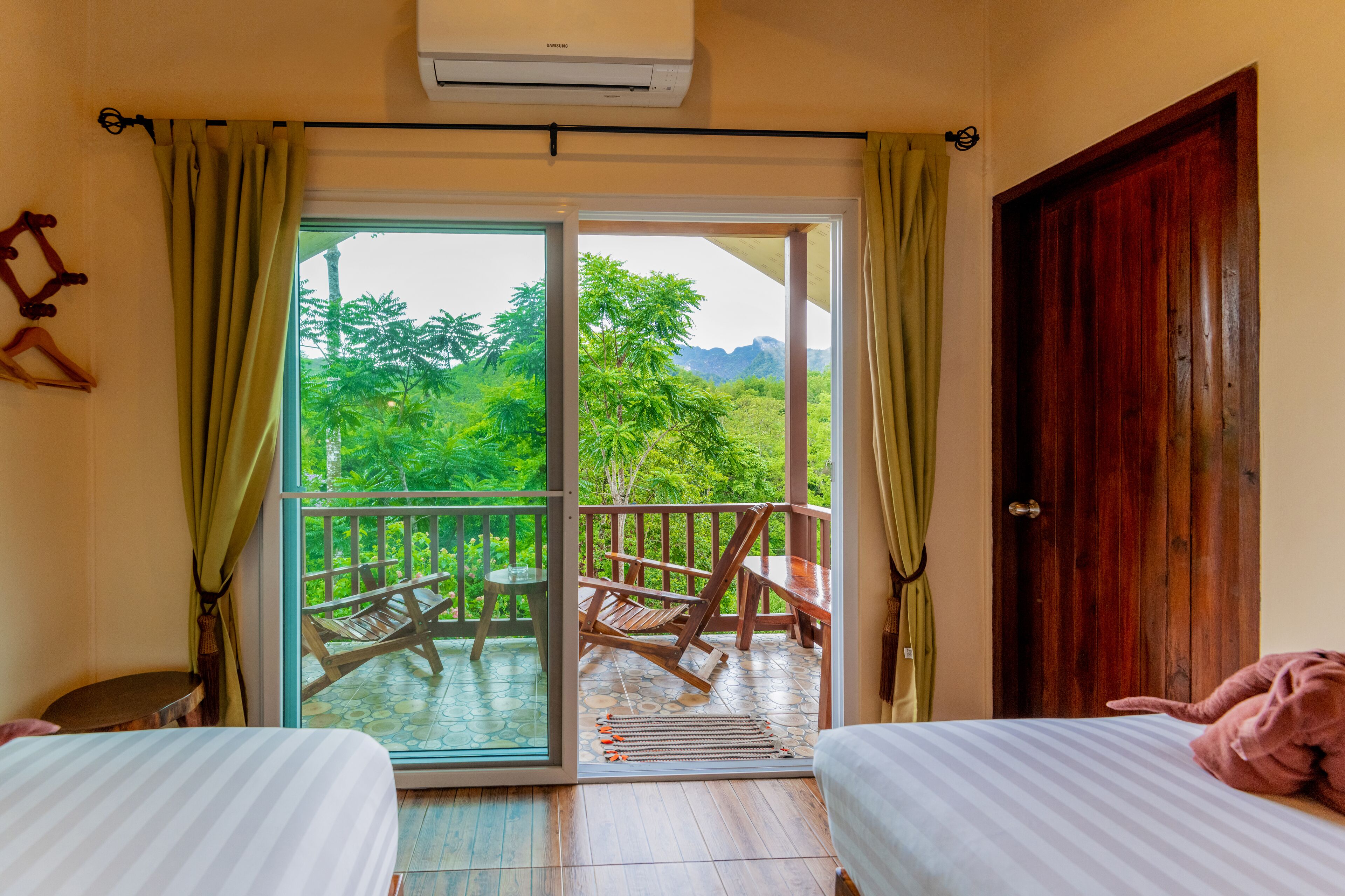 Une chambre avec deux lits, un balcon doté d’une table et de chaises, et une vue sur la verdure environnante.