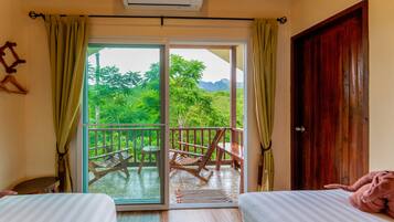A room with two beds, a balcony with a table and chairs, and a view of greenery outside.