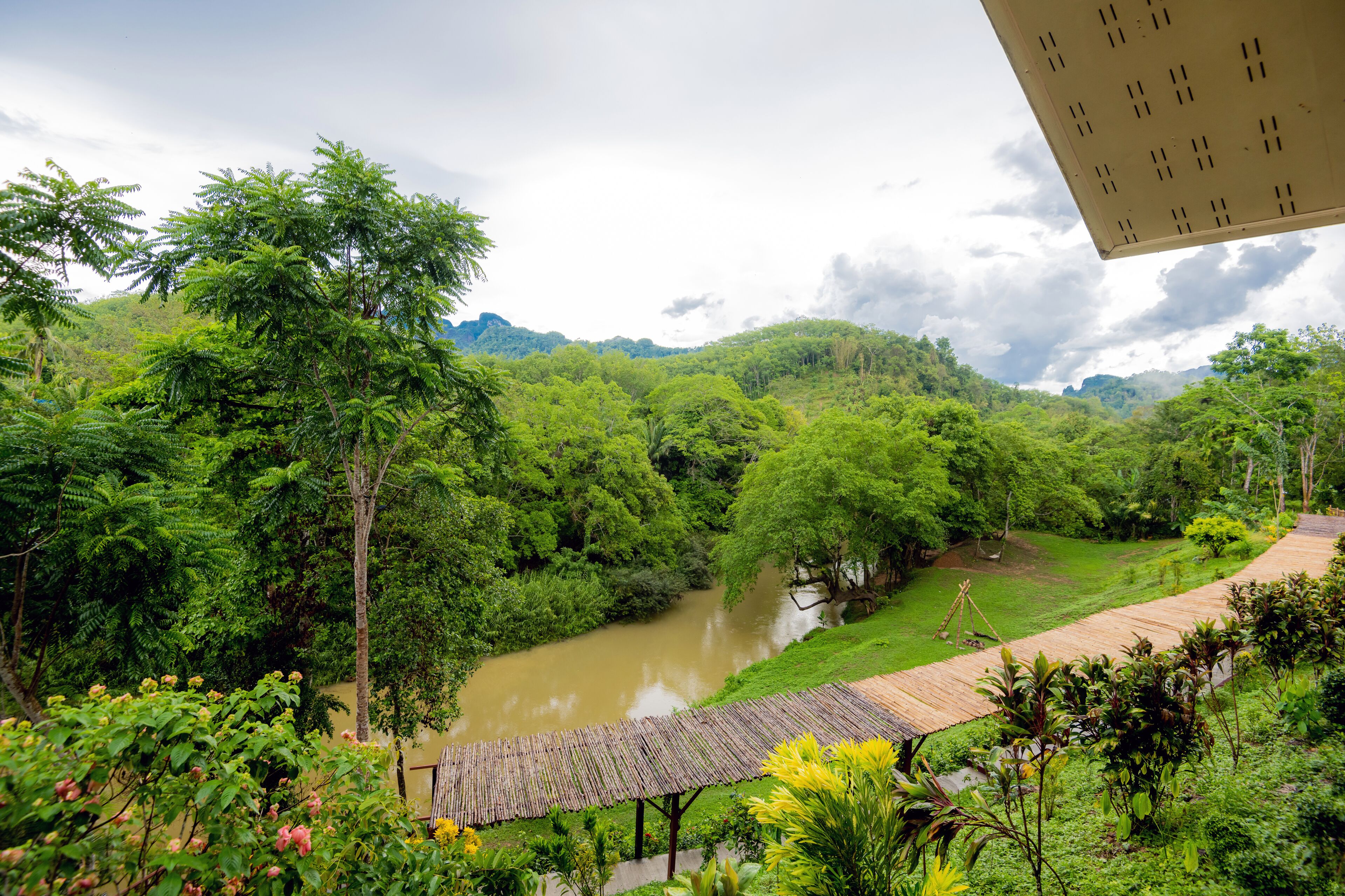 A lush green landscape with a river, a wooden bridge, and a thatched-roof structure.