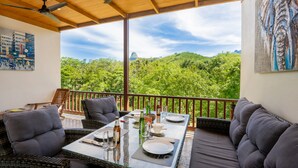 Un área de estar al aire libre techada con una mesa lista para comer, con vistas a un paisaje verde exuberante y una montaña destacada en la distancia.