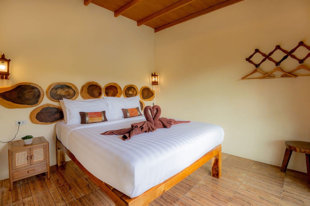 Een net opgemaakt bed met een witte deken en bruine handdoeken, een houten hoofdbord met decoratieve borden, een klein nachtkastje met een plant, en een wandrek met kledinghangers.