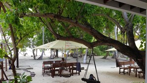 A beachside seating area with wooden benches and tables, a swing, and umbrellas providing shade.