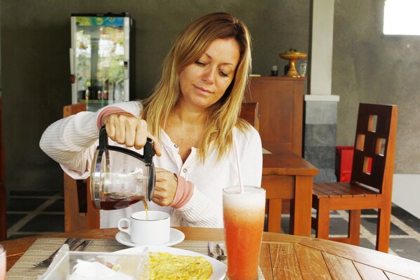 Uma pessoa servindo uma bebida em uma xícara em uma mesa de madeira, com uma bandeja de comida e um copo de chá gelado.