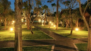 A pathway lined with trees and illuminated by street lamps, leading to a building with a thatched roof. - D' Padang (Pecatu)