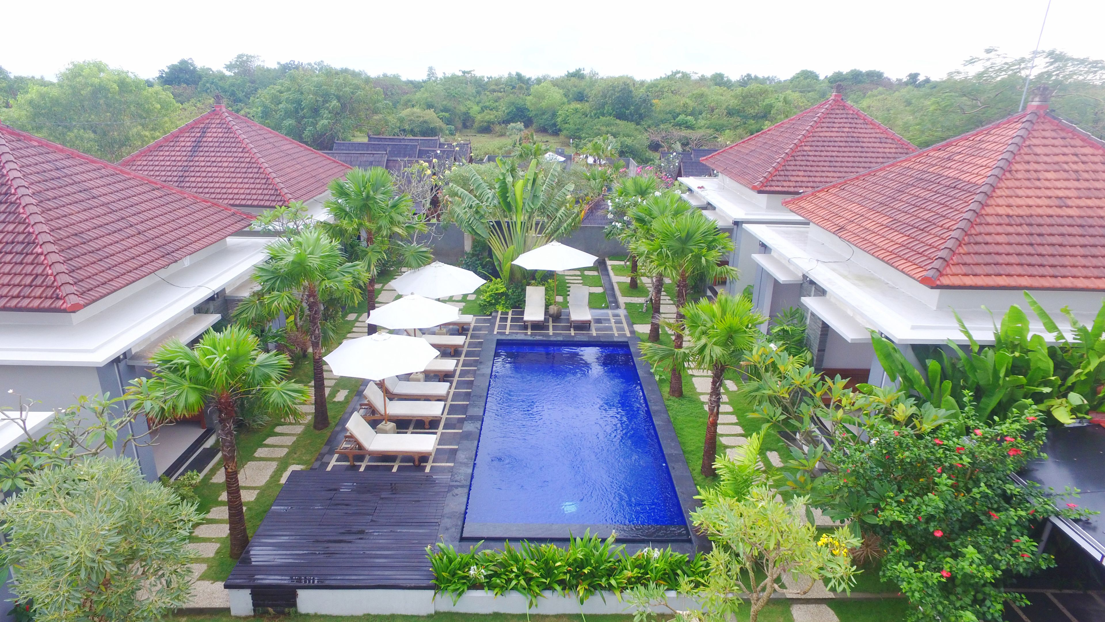 A swimming pool surrounded by lounge chairs and umbrellas, with red-tiled roofs and lush greenery.