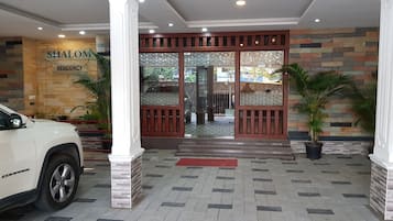 L’entrée d’un hôtel, avec une voiture blanche garée devant, une enseigne indiquant « SHALOM RESIDENCY » et un sol carrelé orné d’un motif.